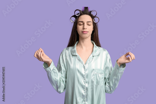 Young woman in pajamas and hair curlers meditating on lilac background