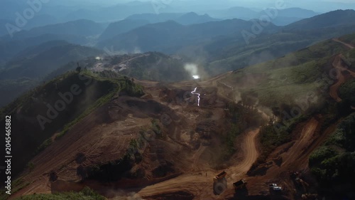 Top-down aerial tracking shot of an active iron ore mine, Red laterite soil and mining equipment contrast with surrounding green forest and mountains in Xieng Khouang province, Lao PDR.