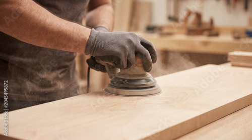 Skilled hands with gloves operate an orbital sander, sanding wood in a workshop. Woodworking detail
