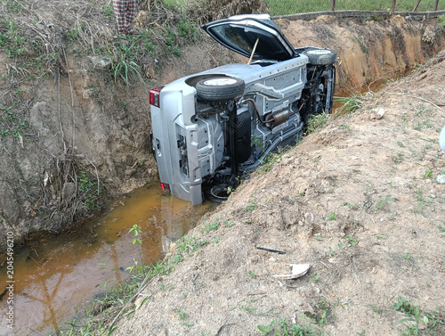 A silver car lies on its side in a muddy ditch beside a dirt road, with the hood open. It appears to be a rural accident scene in malaysia with no visible responders.