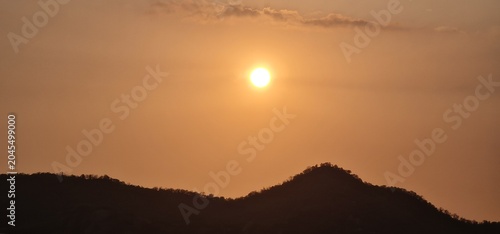 Bright Glowing Sun Setting Over Dark Silhouette of Mountain Ridge in Golden Sky