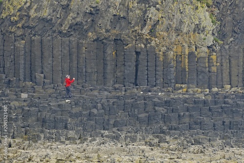 Person with red jacket standing in front of impressive basalt columns on a rocky cliff, Staffa, Mendelsons Hebrides Overture, Inner Hebrides, Scotland, United Kingdom