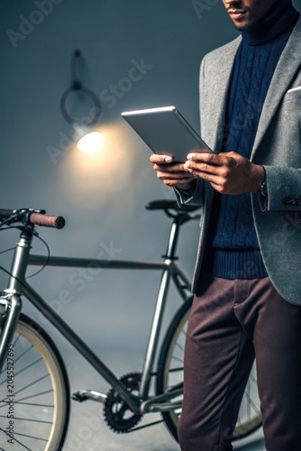 Bike business owner concept. Man in stylish outfit using digital tablet next to a modern bicycle indoors under soft lighting.