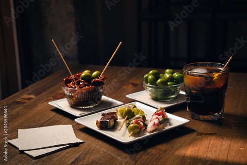 Assorted spanish tapas, including pintxos and olives, arranged on small plates with a vermouth cocktail, creating an inviting scene for socializing and dining