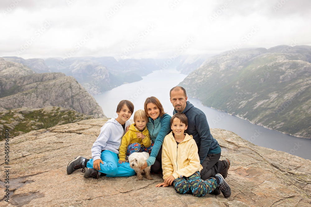 Fototapeta premium Family, enjoying the hike to Preikestolen, the Pulpit Rock in Lysebotn, Norway on a rainy day, toddler climbing with his pet dog the one of the most scenic fjords