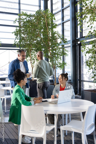 Diverse coworkers wearing business attire meeting around round table in office atrium with laptop