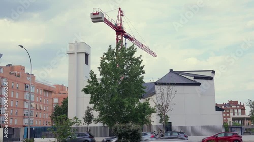 A daytime wide shot of a modern white Catholic parish church building under construction in Madrid, Spain. A large red tower crane stands behind the contemporary church structure and bell tower under 