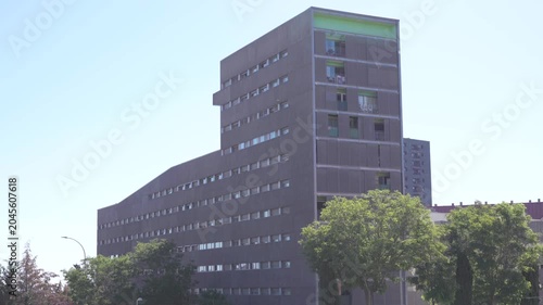A daytime wide shot of a contemporary multi-story residential apartment block with a dark facade and light green accents on the upper balconies in Madrid, Spain. In the foreground, several lush green 