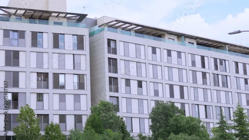 A daytime wide shot of the exterior facade of a contemporary multi-story residential apartment building in Madrid, Spain. The building features large windows and grey panels, partially framed by green