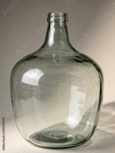 A vintage glass demijohn bottle with a clear, slightly greenish hue. The bottle features noticeable imperfections and a wavy surface texture, typical of hand-blown glass. 