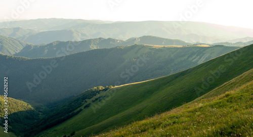 Mountain landscape with lush green slopes, forested hills and soft sunlight illuminating rolling ridges