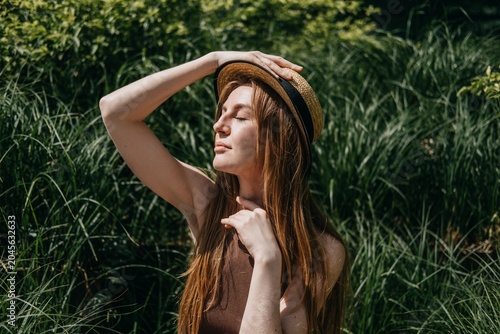 Woman sits on outdoor bench surrounded by green plants holding hand to straw hat. Managing urban heat stress, body hydration techniques , essential sun safe behaviors, summer fatigue prevention