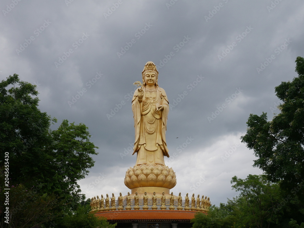 Fototapeta premium Large golden Guanyin statue standing on a lotus pedestal under dramatic storm clouds