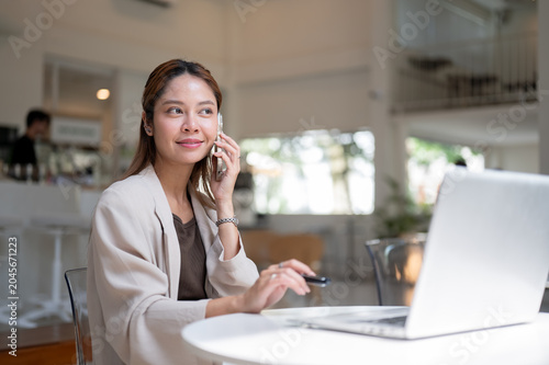 Smiling asian woman holding pen listening to phone call in front of laptop sitting at table in cafe.