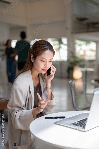 Pretty asian woman holding phone talking on call looking at a laptop while sitting at table in cafe.