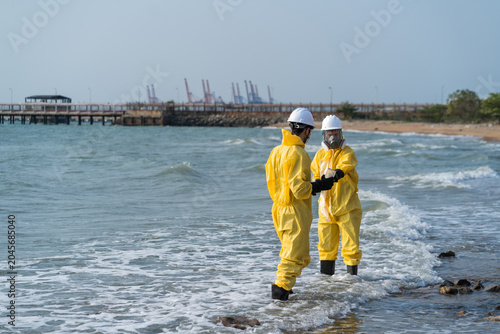 Environmental engineers in hazmat suits collecting and inspecting water samples near industrial gas storage tanks for pollution and safety monitoring.