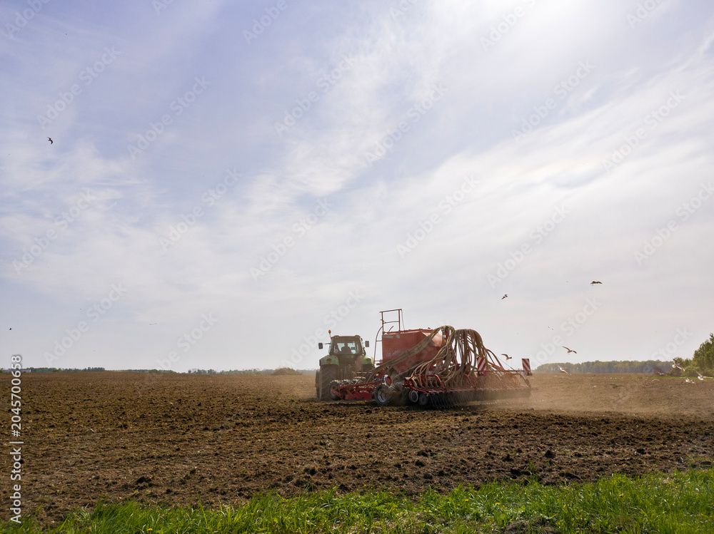 Fototapeta premium Tractor pulling a seeder machine across a field. Farmer planting seed in soil during spring season. Modern agricultural equipment