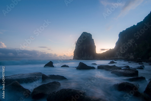 Slika na platnu Serene long-exposure seascape of a monolithic sea stack at Pangasan Beach, Pacitan, with misty waves flowing around foreground boulders at sunset