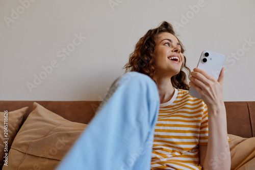 Fotografia Joyful woman relaxing at home on a sofa, looking at a smartphone, striped top, s