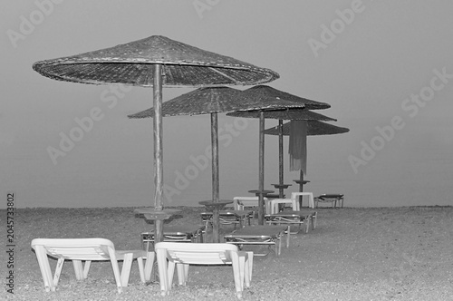 A dramatic black and white fine art photography of a neat row of thatched straw umbrellas and sunbeds. monochrome view.
