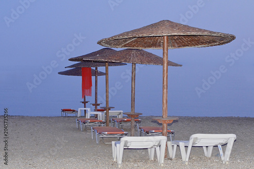 A serene beach scene just after sunset, featuring a neat row of thatched straw umbrellas and sunbeds creating a vanishing point perspective towards the calm sea during dusk.