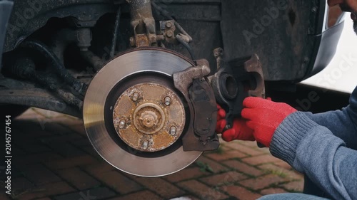 Removing brake caliper assembly from car disc brake system. Repairing vehicle braking mechanism with hands detaching caliper from rotor hub outdoors. Automotive maintenance process focusing on brake