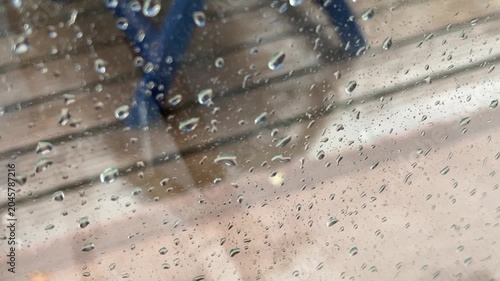 Close-up shot of raindrops on a window pane with a blurred background showing the wet wooden decking and outdoor rug of a balcony during a heavy rainstorm.