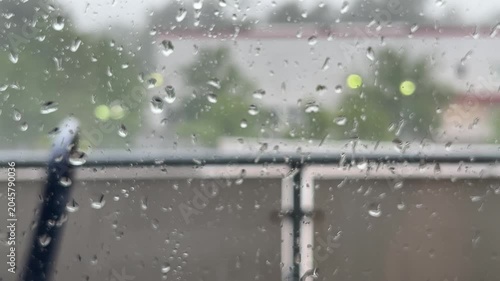 Close-up shot of raindrops collecting on a window pane during a heavy storm, with a blurry background of trees and buildings under a grey, overcast sky.