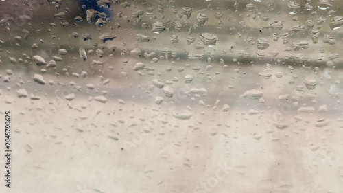 Close-up shot of raindrops on a window pane with a blurred background showing the wet wooden decking and outdoor rug of a balcony during a heavy rainstorm.