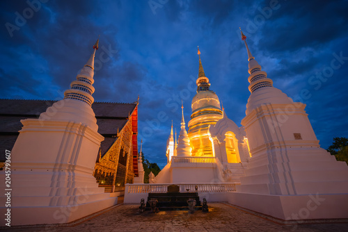 The golden pagoda of Wat Suan Dok temple illuminated at twilight on a rainy season night with a Buddhist monk standing by the wall in Chiang Mai, Thailand