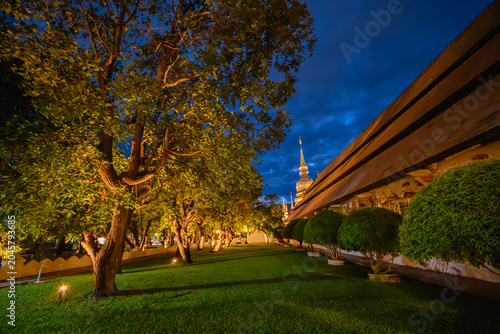 The golden pagoda of Wat Suan Dok temple illuminated at twilight on a rainy season night with a Buddhist monk standing by the wall in Chiang Mai, Thailand