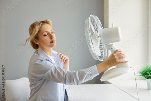 Obraz na plátne Portrait of young upset woman sitting at the desk at home and suffering from summer heat in front of fan