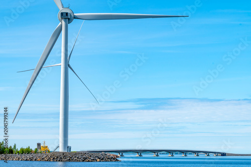 Giant wind turbine standing near the modern Hailuoto bridge in Finland, alternative energy source infrastructure and sea road connection in Bothnian Bay under a blue sky.
