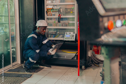 Black male maintenance specialist in safety gear tracking logistics data or automation software on a tablet screen during factory operation, showcasing digital transformation and workforce diversity.
