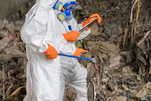 Photos Hazardous Waste Management:Two specialists in a full hazmat suit,respirator examines environmental samples amidst a large pile of refuse, highlighting critical hazardous waste management and safety