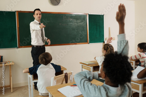 Description (EN): Professional Caucasian schoolteacher explaining a math formula on a blackboard while young multiethnic children actively listening, highlighting primary school curriculum and modern 
