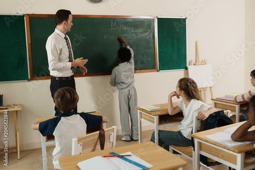Description (EN): Professional Caucasian schoolteacher explaining a math formula on a blackboard while young multiethnic children actively listening, highlighting primary school curriculum and modern 
