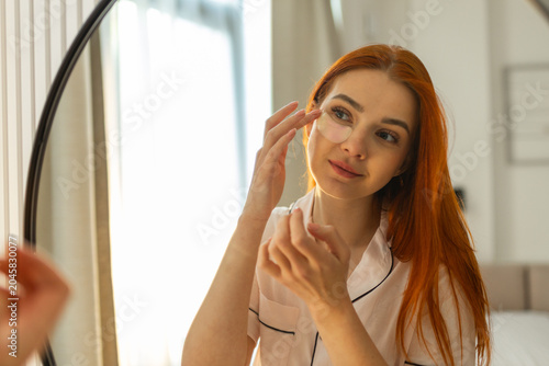 Young woman applying hydrogel eye patches while checking her reflection in mirror