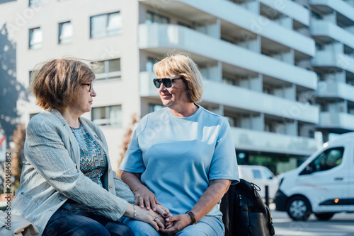 Fotografie Senior Woman And Midlife Friend Holding Hands On City Bench