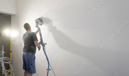 Professional contractor polishing drywall surface with electric wall sander and integrated vacuum system, creating smooth finish during residential renovation