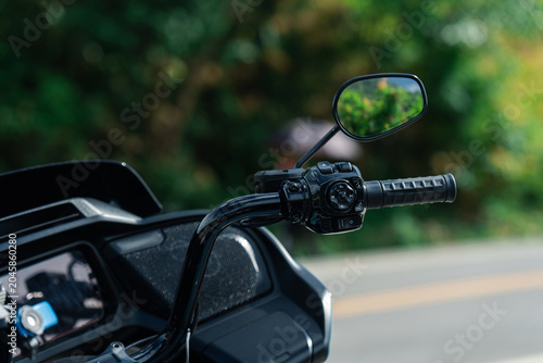 Close up of motorcycle handlebar with mirror, showcasing sleek design and black finish. background is blurred greenery