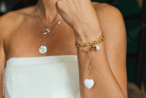 Fotografía Close up of a woman hand wearing a gold chain bracelet with a white heart charm in front of layered silver necklaces