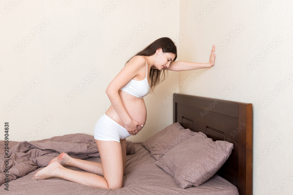 Obraz premium beautiful pregnant woman touching her tummy and keeping one hand on her back at home on bed
