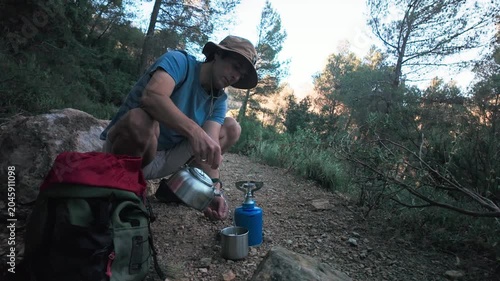 Man camping in forest pouring water from kettle into mug next to backpack and camping stove