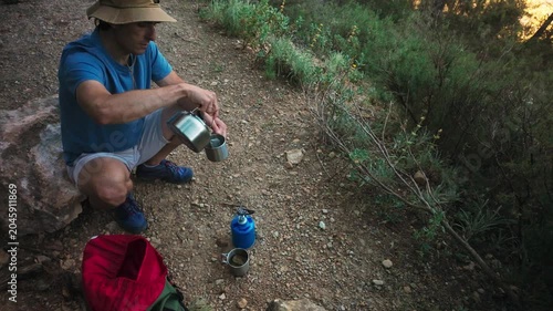Man camping on a rocky trail pouring tea from a kettle into a mug beside backpack and stove