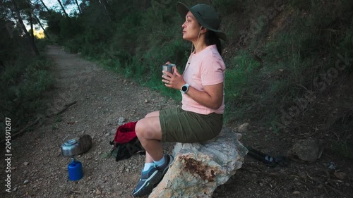 Woman resting on a rock during a solo hike with camping gear and a hot drink in nature