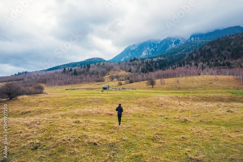 Photos A person walks alone across a lush green meadow, surrounded by gentle hills and towering mountains