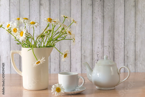 Summer morning still life; white cup of tea or coffee, tea pot and bouquet of white daisy flowers on a table against white wooden background, copy space
