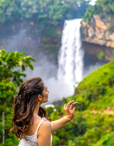 Woman enjoys a blurred waterfall cascading through dense greenery, her hand raised towards the scene
