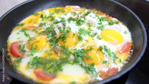 Macro recording of breakfast. Cooking eggs with tomatoes and herbs on the stove in a frying pan at home in the morning.